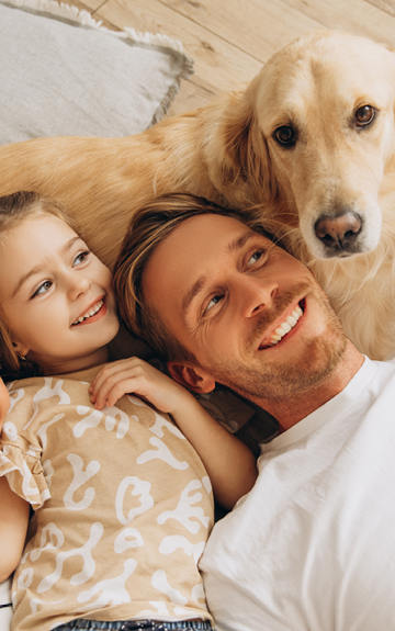 Eine Familie mit Hund liegt auf dem Boden. Vater, Mutter und Tochter lächeln den Hund an.